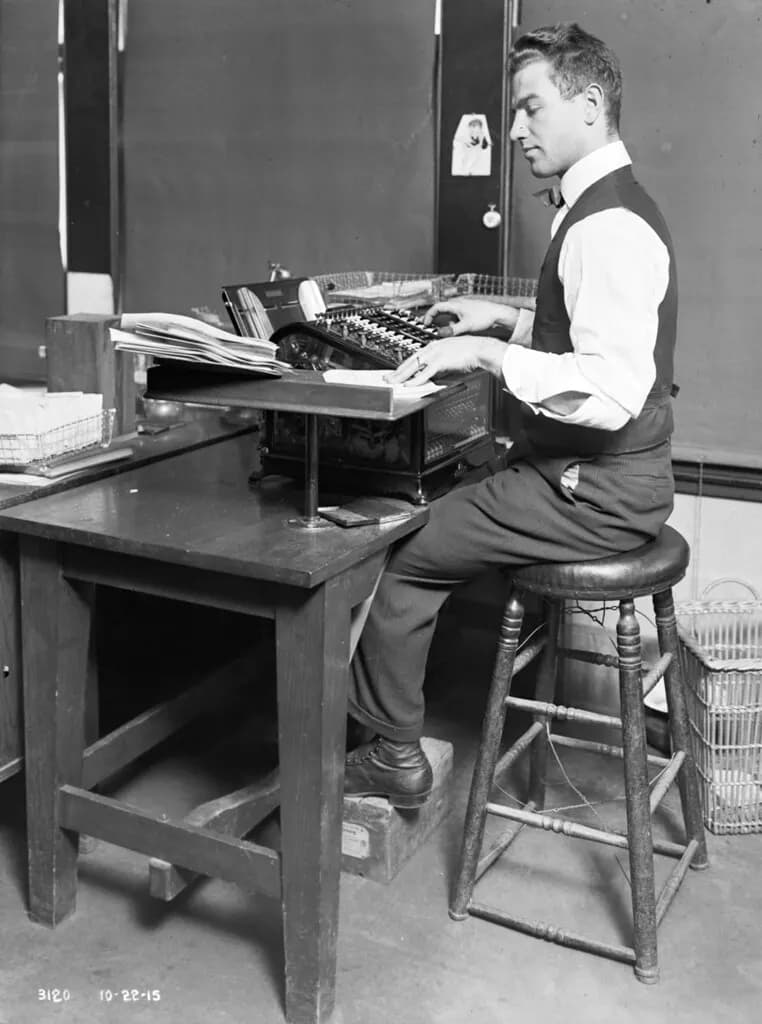 A 1915 municipal clerk working at a mechanical adding machine at a wooden desk