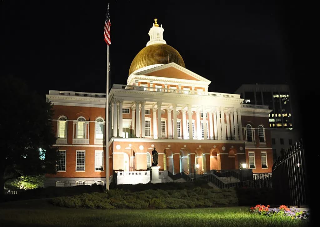 The gold-domed Massachusetts State House in Boston lit up at night behind a lawn and flagpole