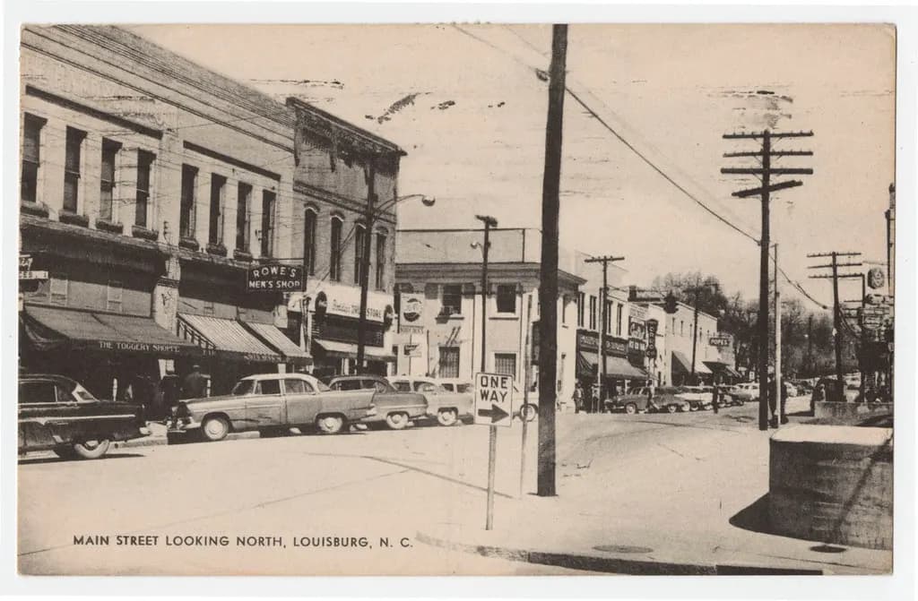 Black-and-white vintage postcard of Main Street in Louisburg, North Carolina, with storefronts, awnings and parked 1950s cars