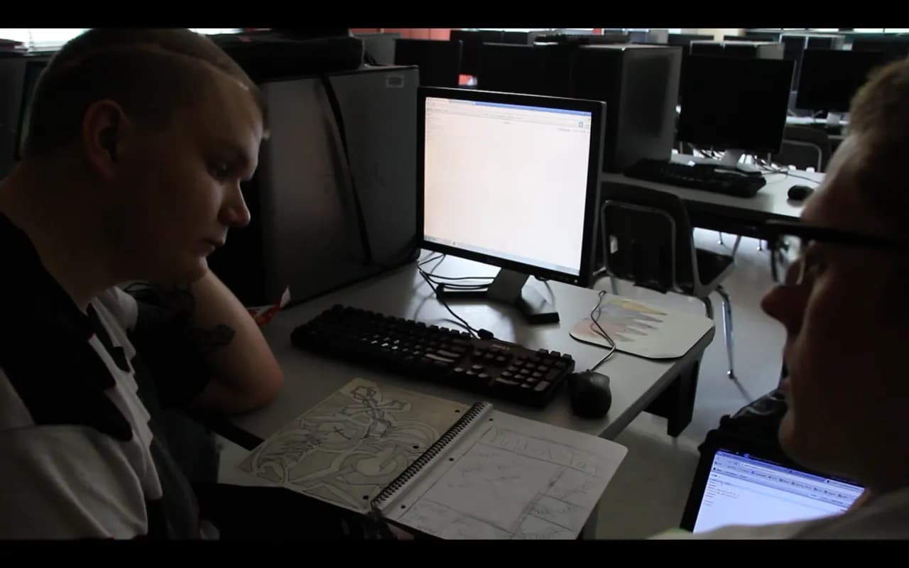 Two people review a paper sketch of a web page layout next to a desktop computer in a computer lab
