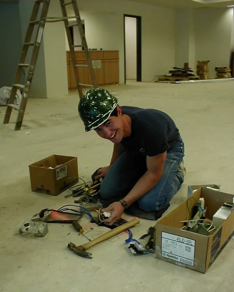 A young electrician in a hard hat kneeling on a job site floor, sorting wiring and tools from cardboard boxes