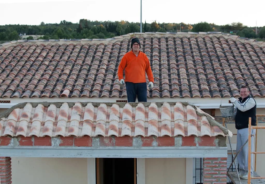 Two workers on a clay tile roof, one standing on the ridge in an orange sweater and one on a ladder below
