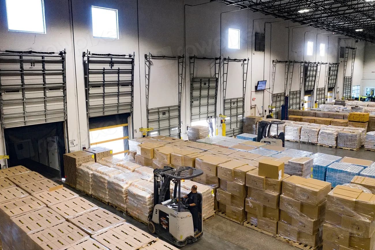 Forklift operator moving pallets of boxed food in a large warehouse with loading dock doors